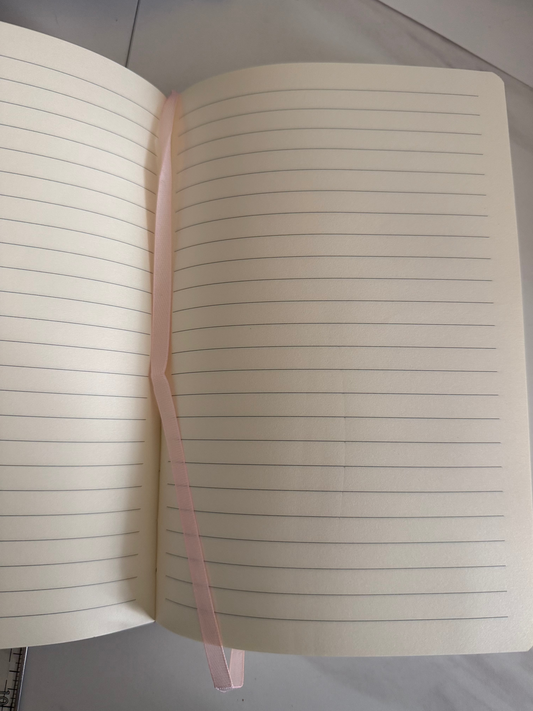 An open pink-colored ruled notebook with a thin pink ribbon bookmark spanning across the pages, photographed from above on a light gray surface.