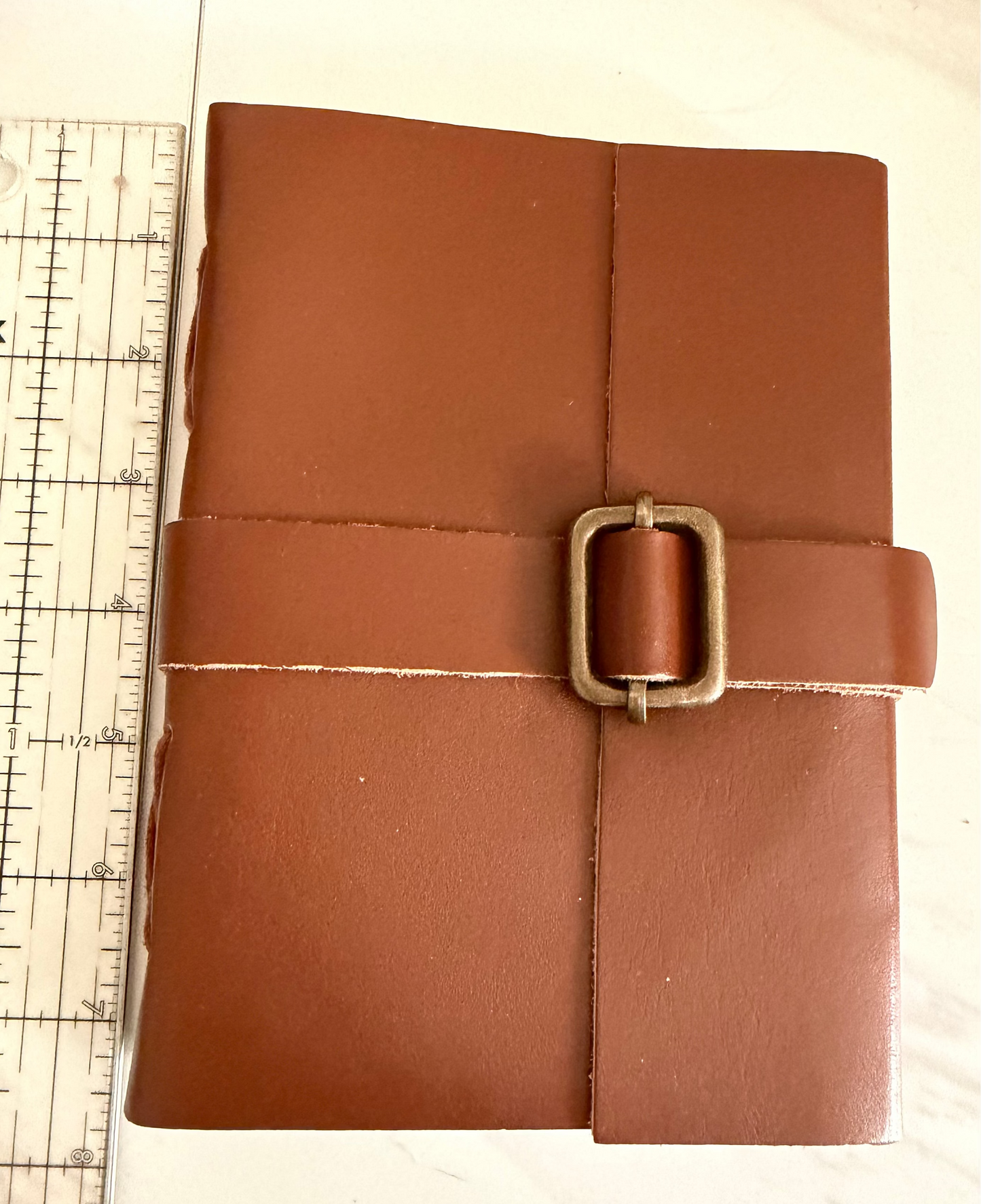 A brown leather journal or notebook with a wrap-around strap and square metal buckle, shown next to a ruler on a light surface.