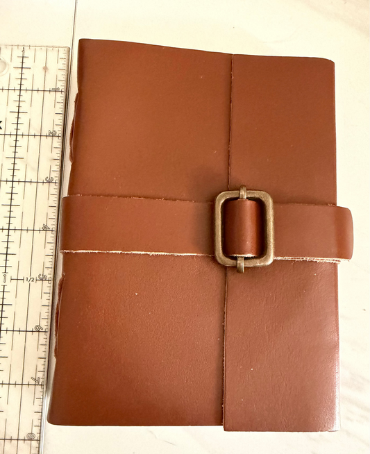 A brown leather journal or notebook with a wrap-around strap and square metal buckle, shown next to a ruler on a light surface.