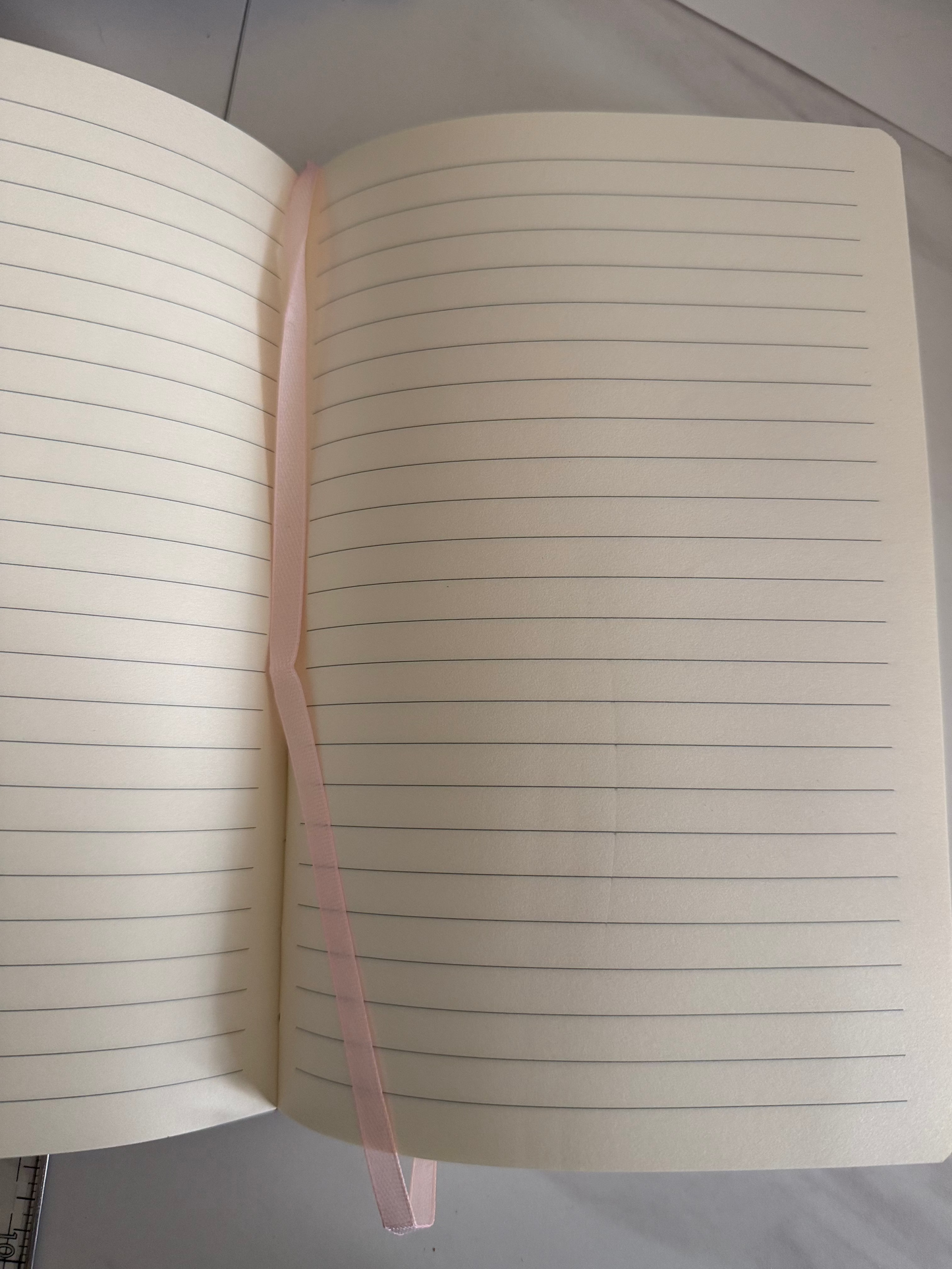An open pink-colored ruled notebook with a thin pink ribbon bookmark spanning across the pages, photographed from above on a light gray surface.