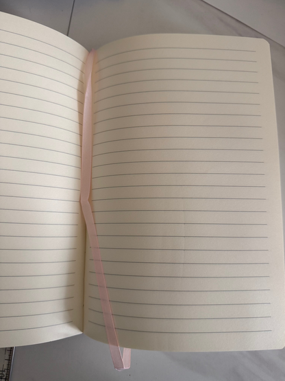 An open pink-colored ruled notebook with a thin pink ribbon bookmark spanning across the pages, photographed from above on a light gray surface.