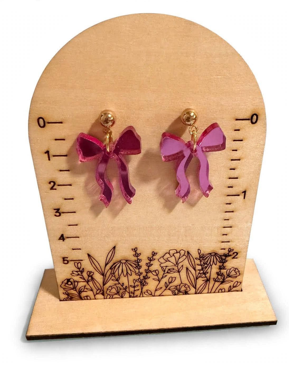 A pair of magenta pink bow-shaped stud earrings with gold posts, displayed on a wooden arch-shaped measurement stand with floral engraving.