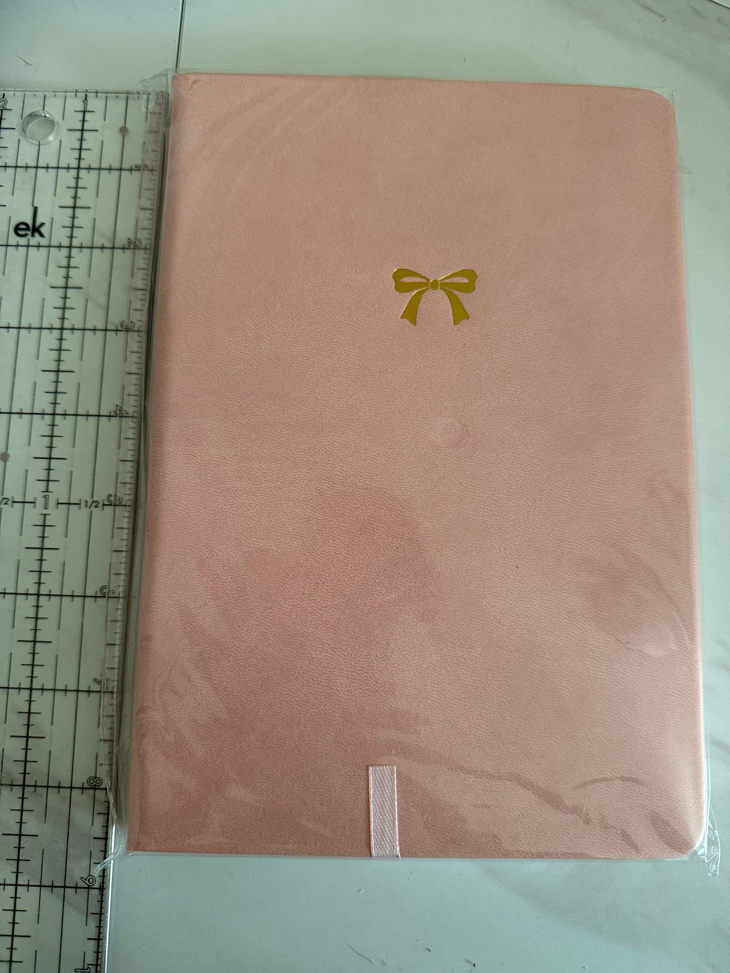 A pink rectangular item wrapped in clear plastic, with a small gold bow-like emblem near the lower right. A clear ruler is placed along the top edge for scale on a light surface.