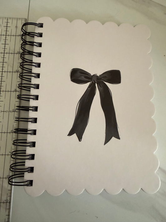 A white scalloped-edge notepad with a black spiral binding, featuring a black ribbon bow illustration in the center, shown with a ruler at the top for scale.